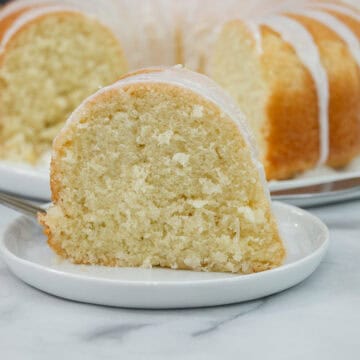 Slice of coconut pound cake on a white plate with the full bundt cake in the background.