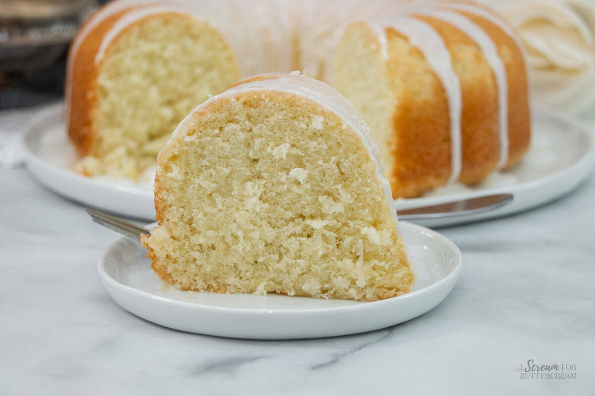 Slice of coconut pound cake on a white plate with the full bundt cake in the background.