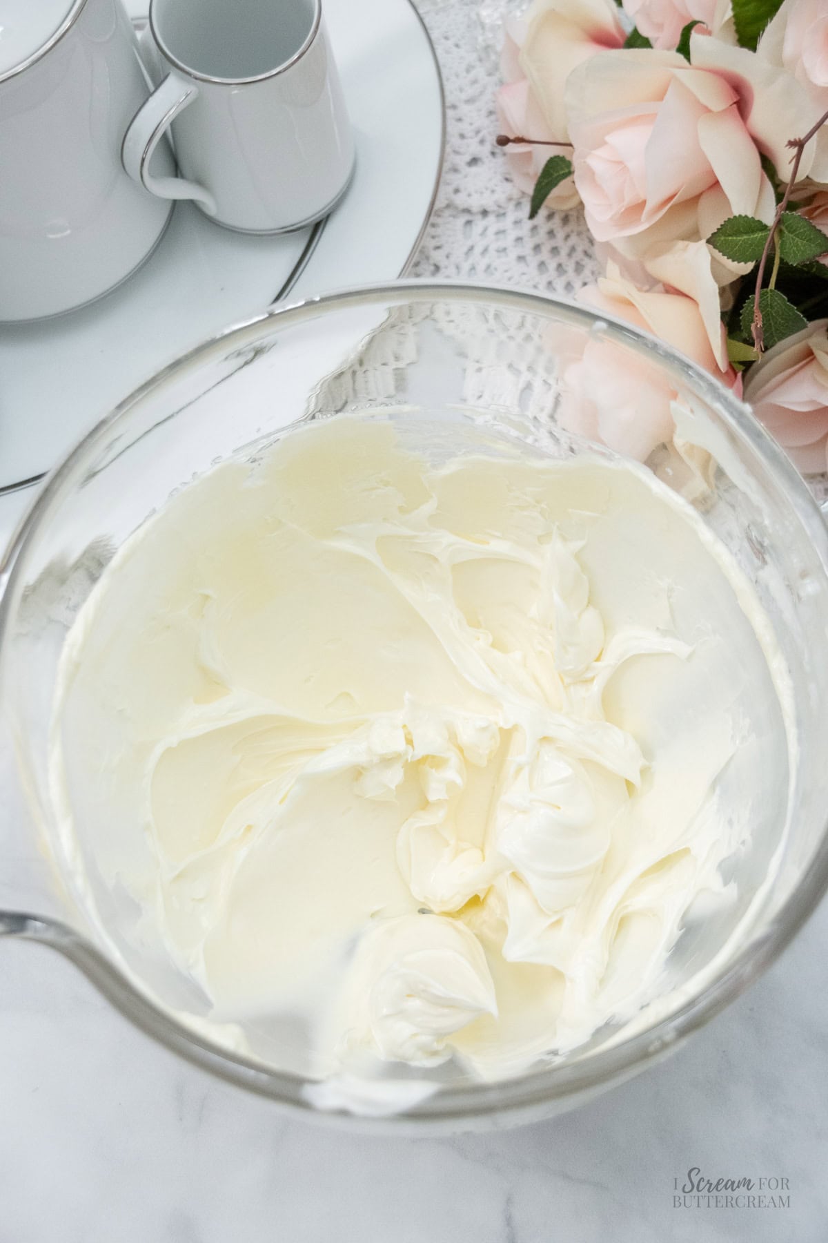 Glass bowl filled with creamy wedding cake frosting, styled beside a tea set and soft pink roses on a lace tablecloth.
