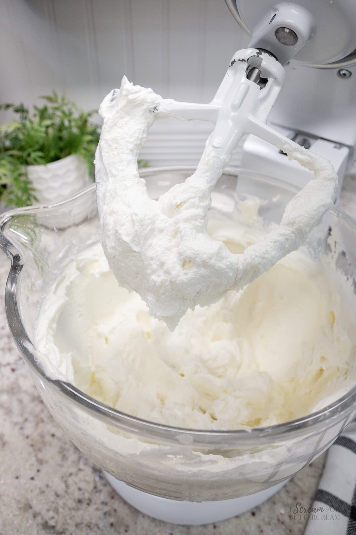 Glass mixing bowl filled with fluffy white frosting on a stand mixer, with frosting clinging to the paddle attachment.