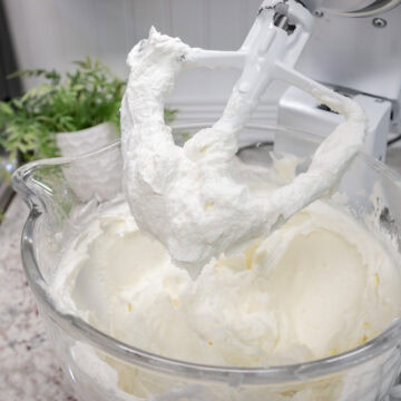 Stand mixer with a glass bowl filled with whipped wedding cake frosting, with fluffy frosting coating the paddle attachment.