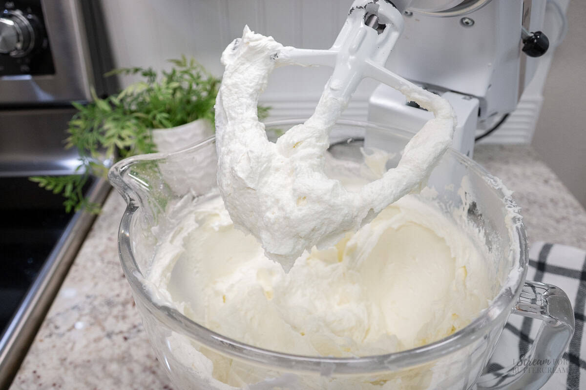 Stand mixer with a glass bowl filled with whipped wedding cake frosting, with fluffy frosting coating the paddle attachment.