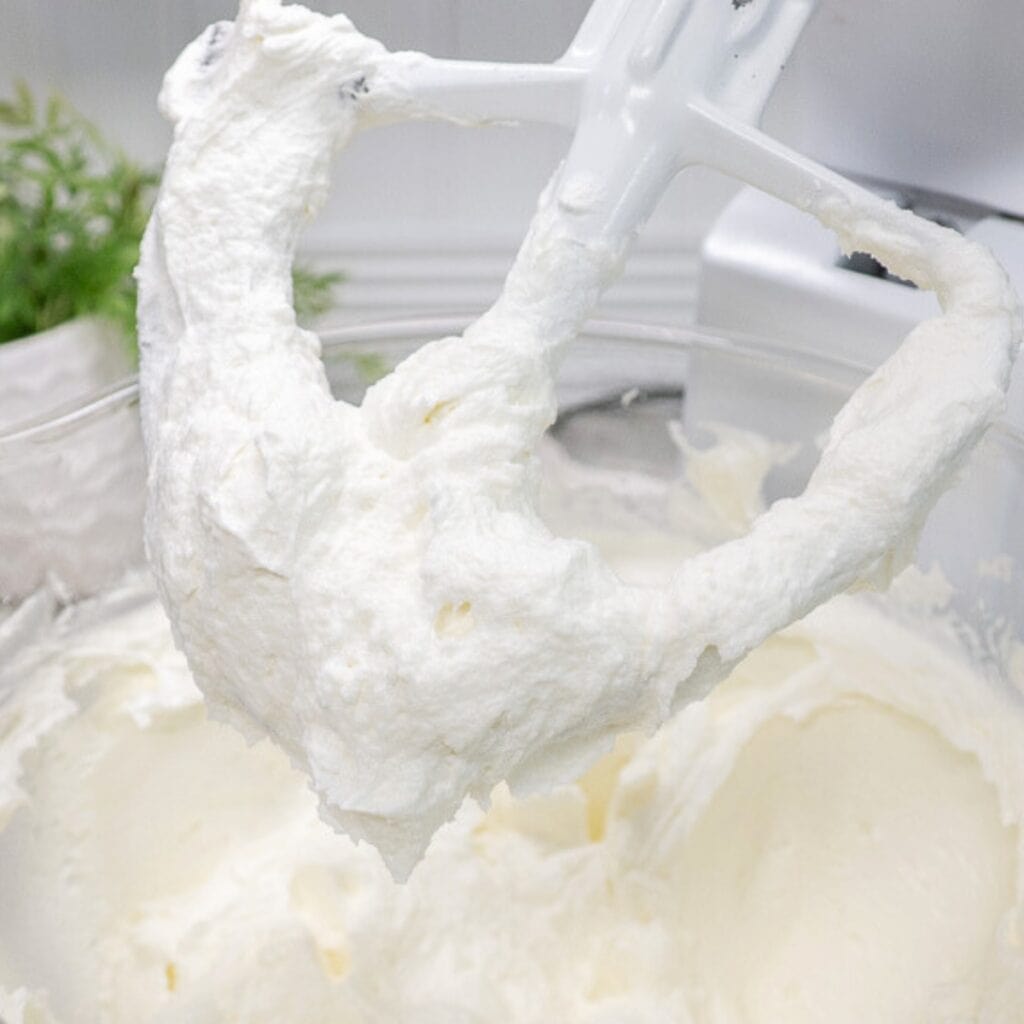 "Close-up of a stand mixer paddle attachment covered in fluffy, whipped buttercream frosting over a mixing bowl.