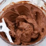 Fluffy chocolate buttercream frosting in a clear mixing bowl with a white paddle attachment, surrounded by white flowers.