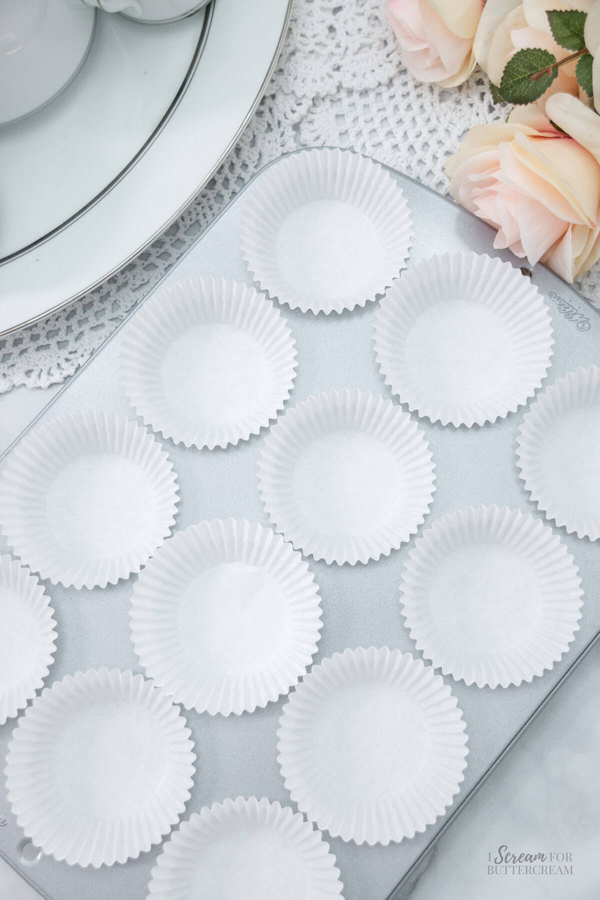 Empty white cupcake liners arranged in a silver muffin tin, set on a lace tablecloth with soft pink roses nearby.