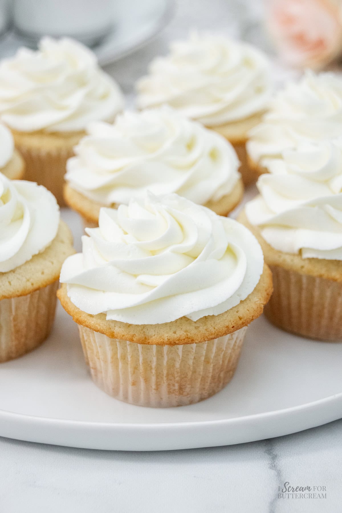 Plate of sour cream white cupcakes topped with piped swirls of creamy white frosting, arranged neatly on a marble surface.