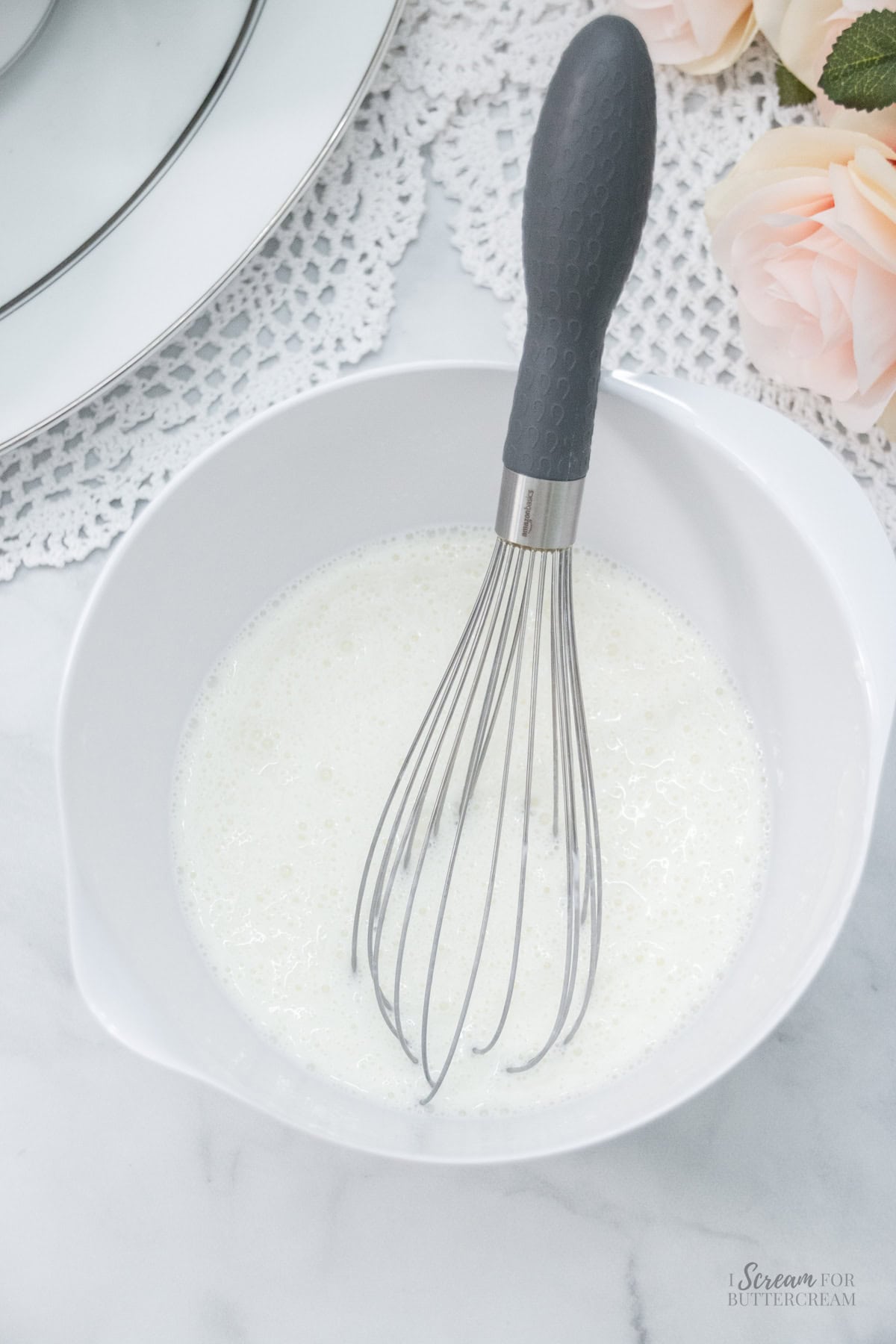White mixing bowl filled with a frothy liquid mixture and a metal whisk, resting on a marble surface with lace and roses nearby.
