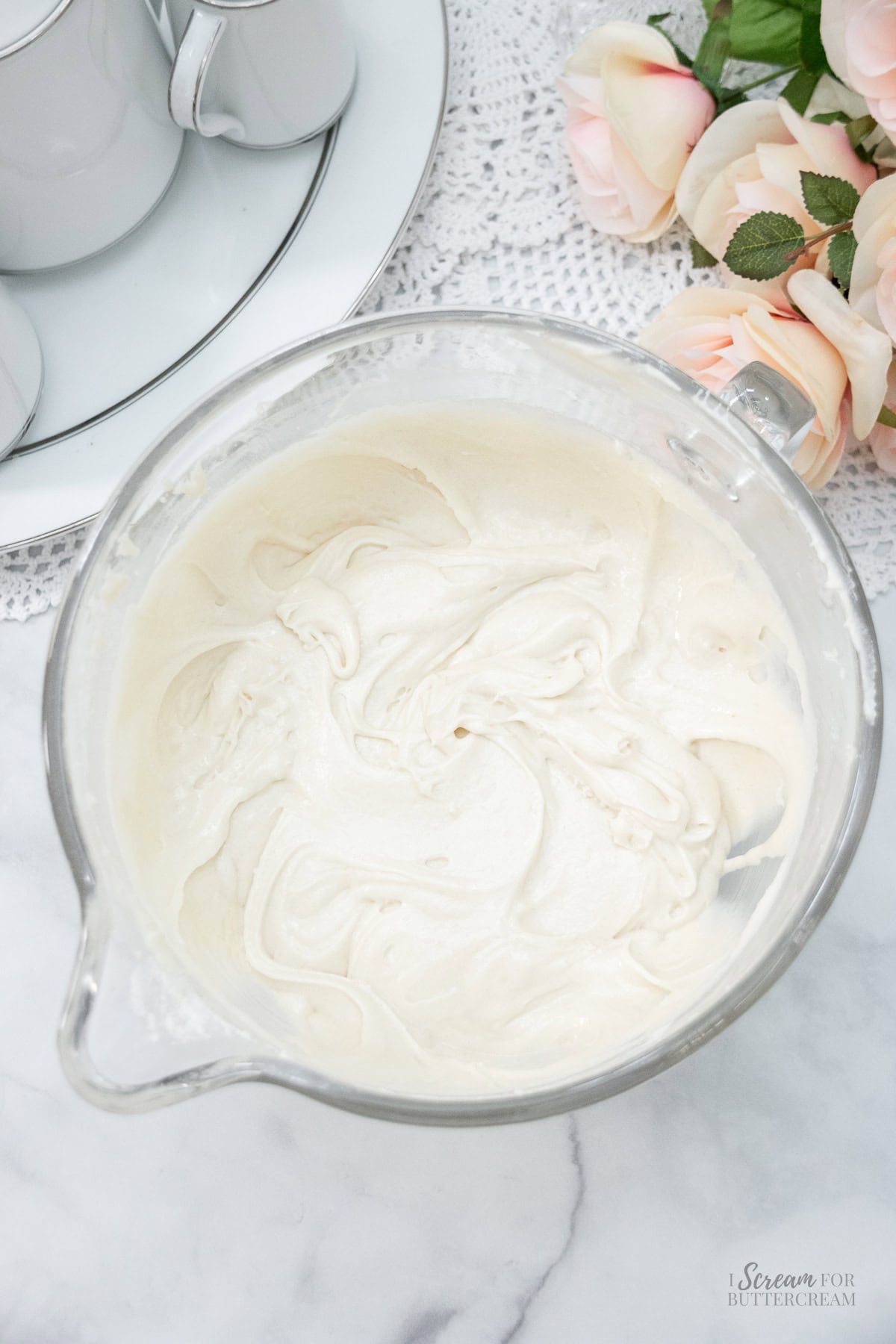 Glass mixing bowl filled with smooth cupcake batter, set on a marble surface with lace and pale pink roses nearby.