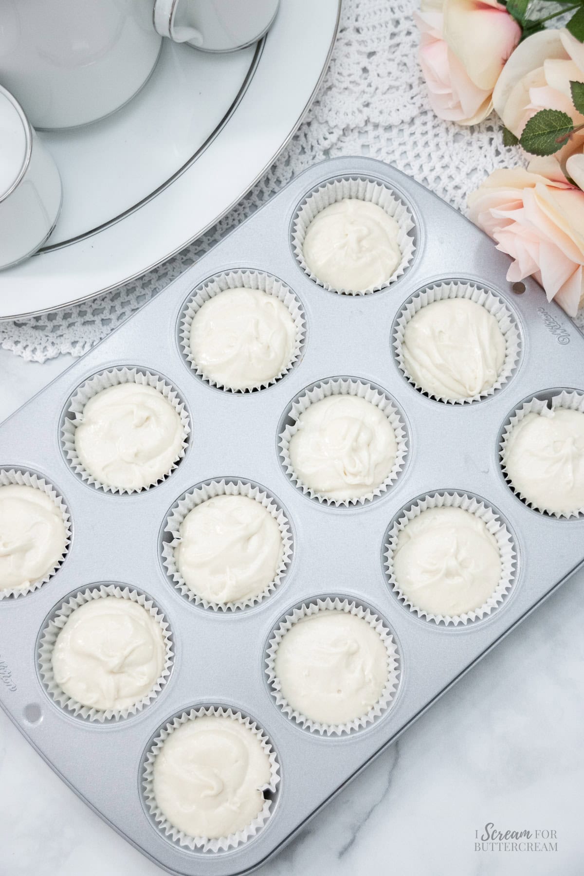 Cupcake pan filled with white paper liners and cupcake batter, ready for baking, set on a marble surface with lace and roses.