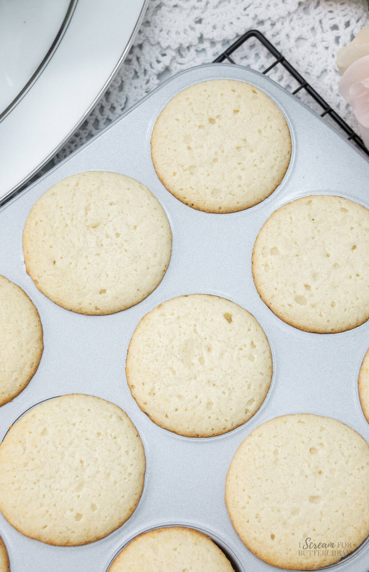 Freshly baked golden cupcakes in a muffin tin, cooling on a wire rack over a lace tablecloth.