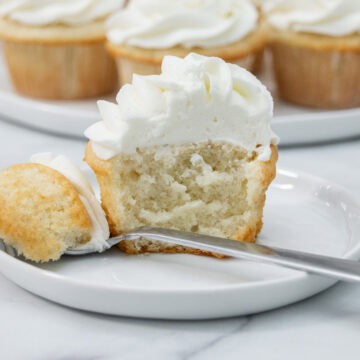 Half of a frosted sour cream white cupcake on a white plate with a fork, surrounded by whole cupcakes in the background.