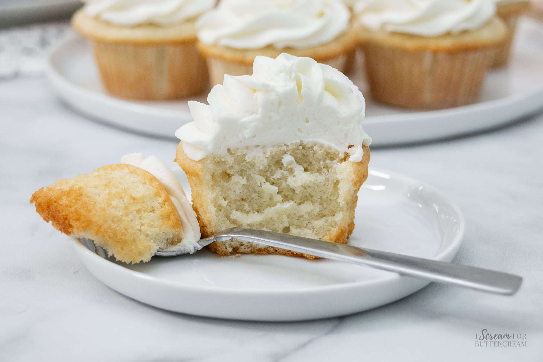 Half of a frosted sour cream white cupcake on a white plate with a fork, surrounded by whole cupcakes in the background.