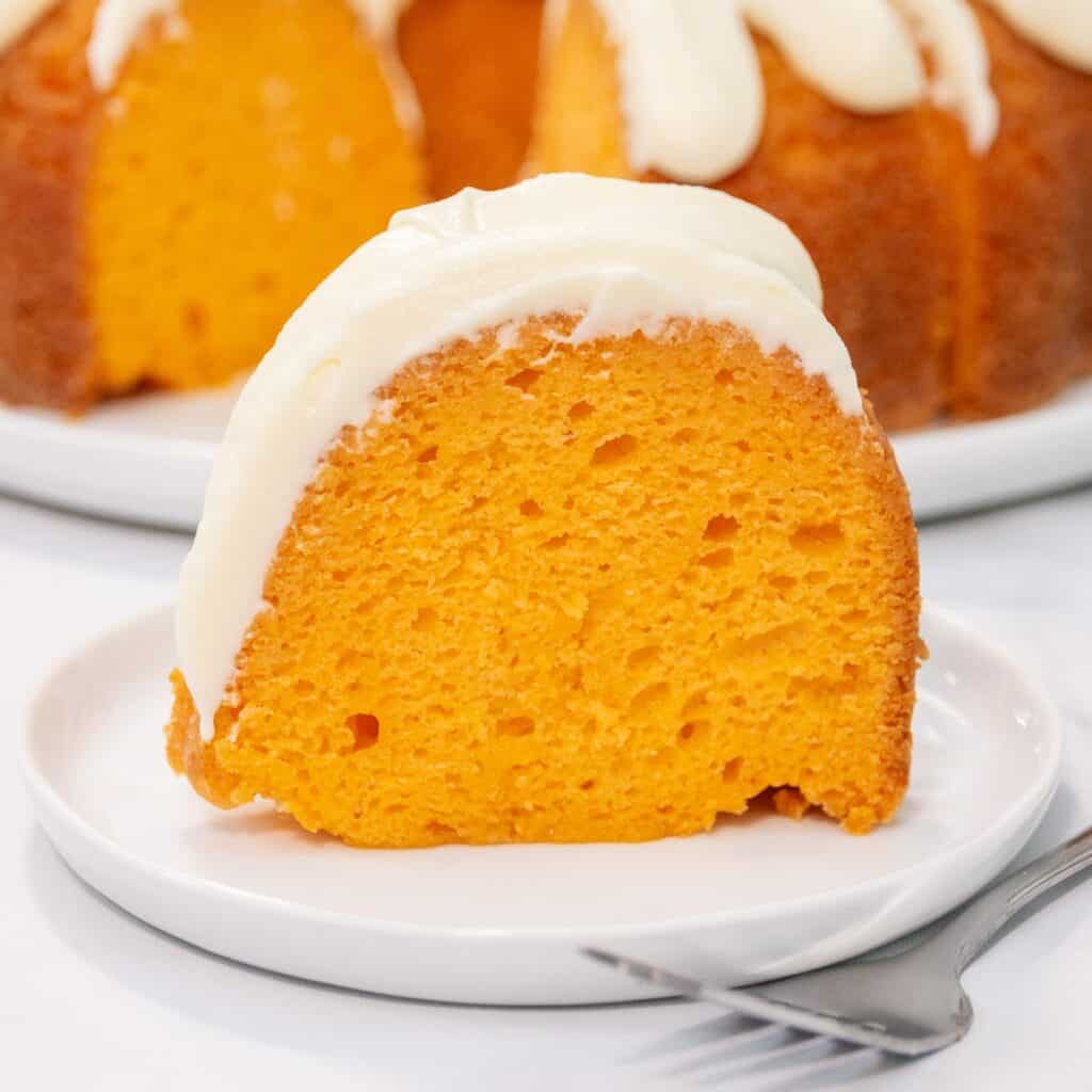A slice of vibrant orange bundt cake with creamy white frosting sits on a white plate with a fork beside it.
