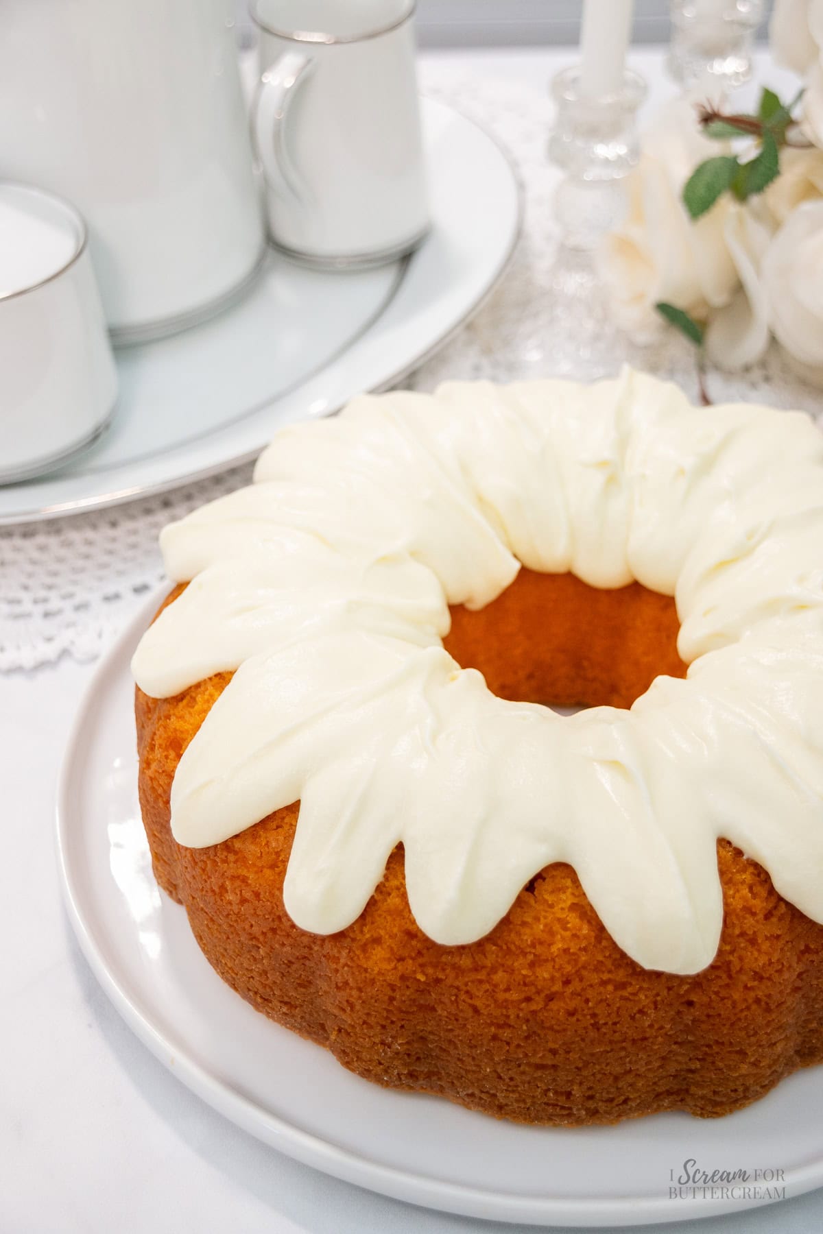 A golden orange bundt cake is topped with thick swirls of cream cheese frosting and displayed on a white plate beside a tea set and roses.