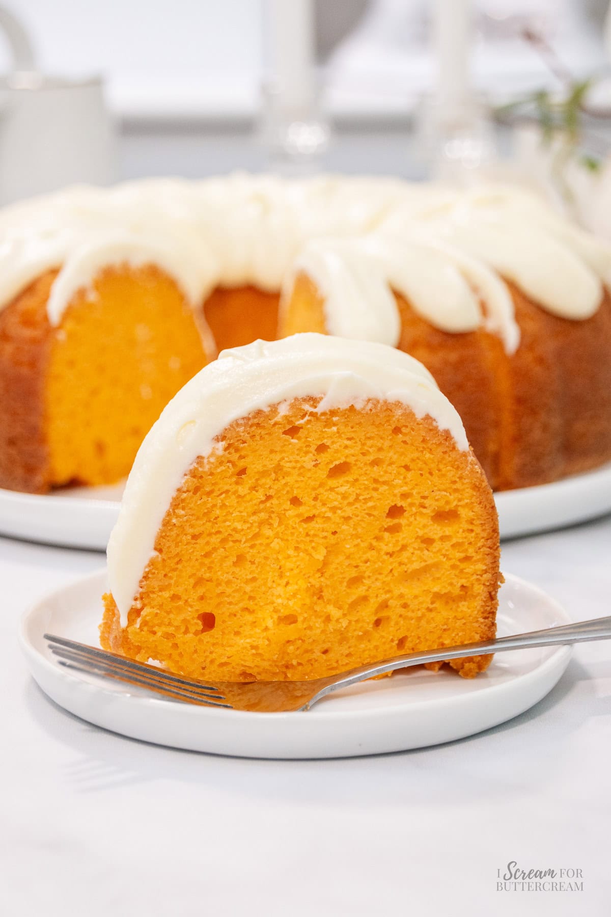 A slice of vibrant orange bundt cake with creamy white frosting sits on a small plate with a fork, in front of the full frosted cake.