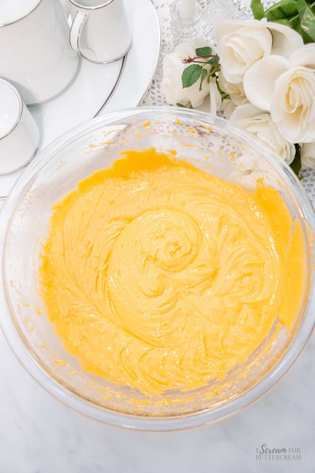 A glass bowl filled with smooth, orange cake batter sits on a marble surface beside a white rose arrangement and a tea set.