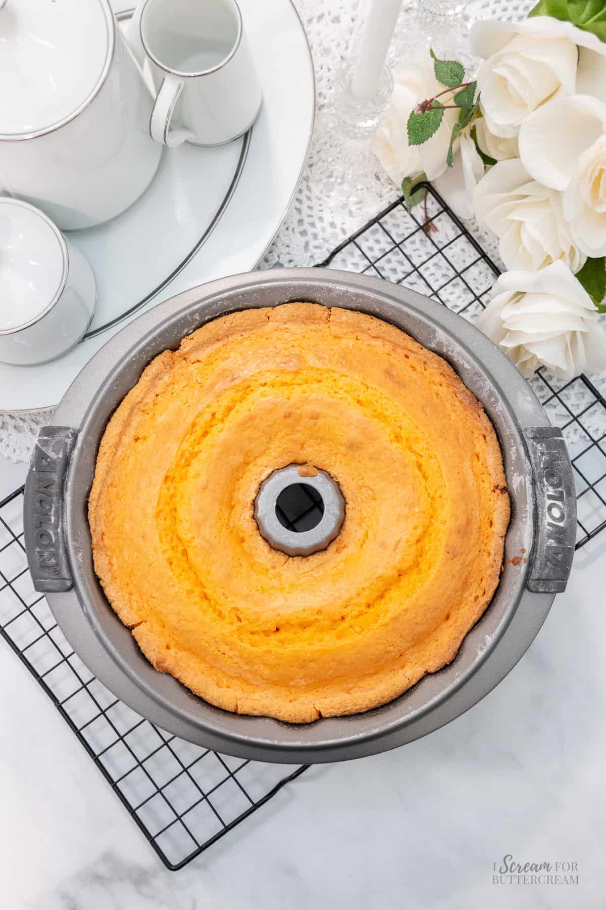 A freshly baked orange bundt cake rests in a pan on a cooling rack beside white roses and a white china tea set.
