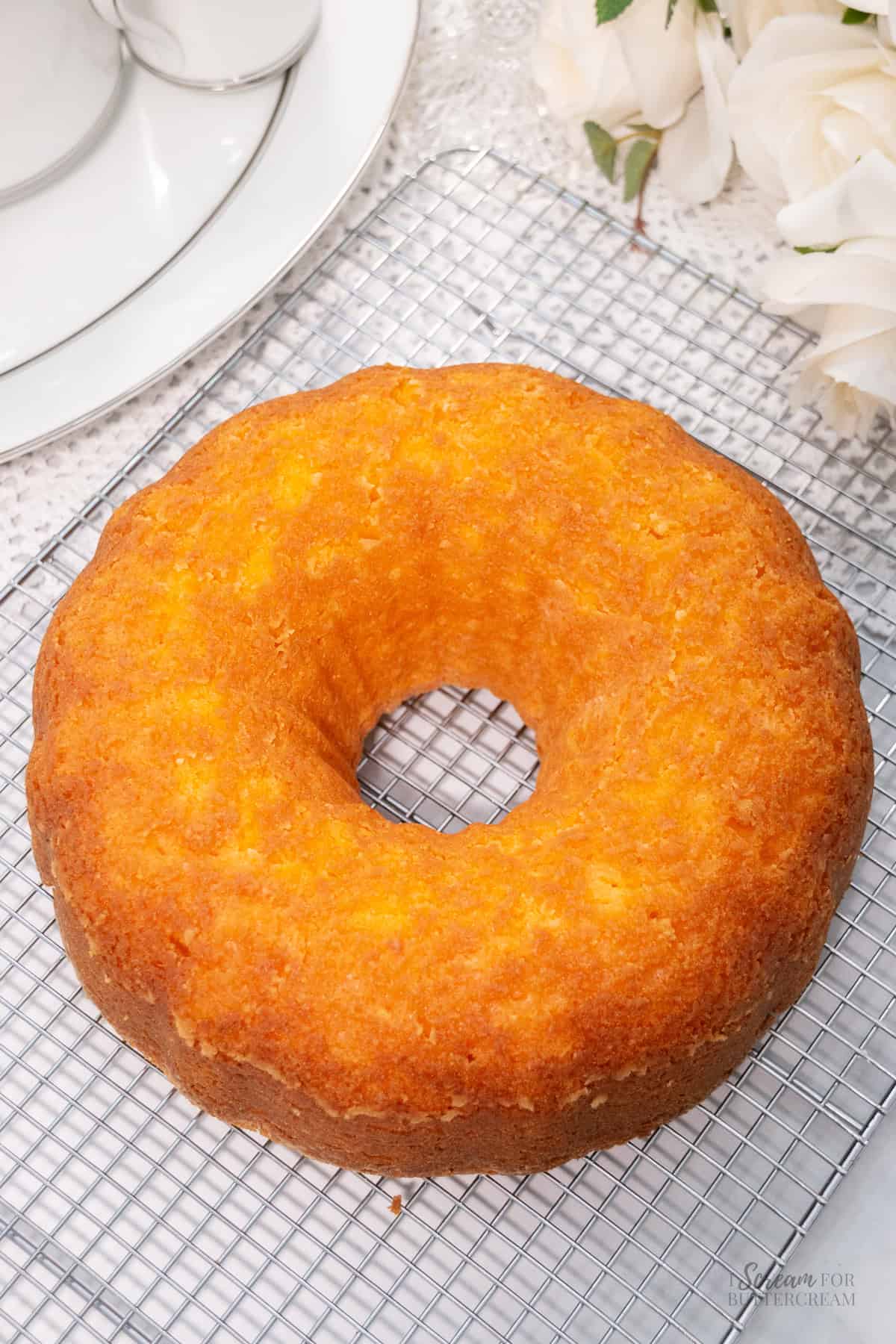 A golden orange bundt cake cools on a wire rack next to a white tea set and a bouquet of cream-colored roses.