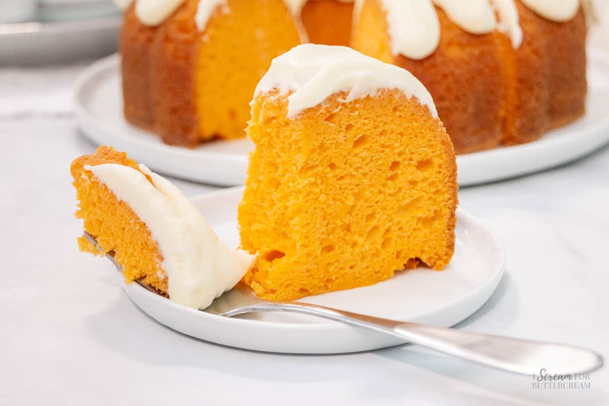 A slice of orange creamsicle bundt cake with white frosting sits on a plate with a fork holding a bite-sized piece in the foreground.