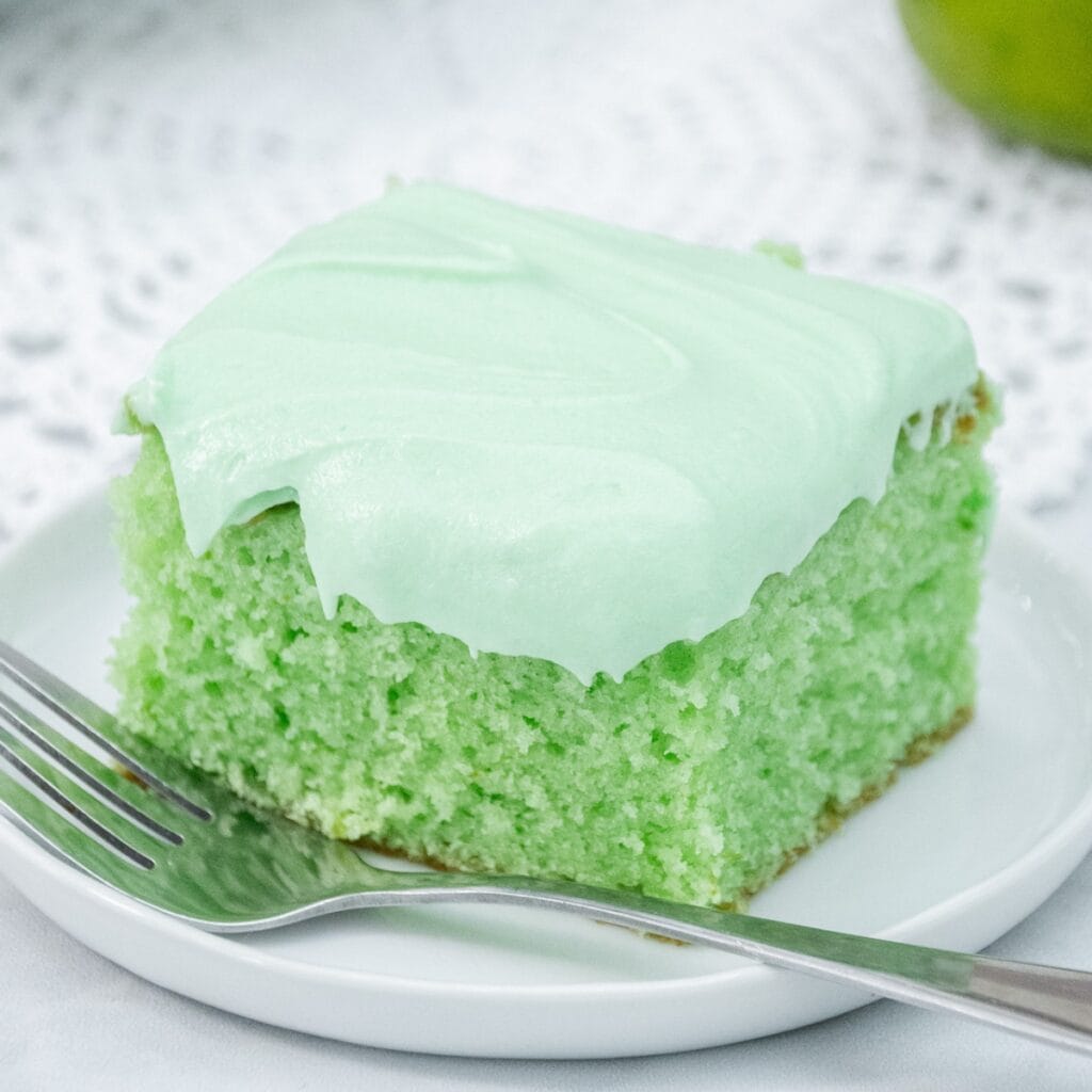 Slice of light green cake with fluffy green frosting on a white plate, served with a fork.