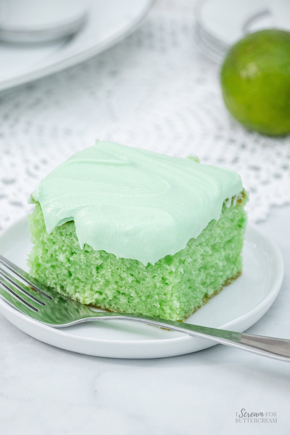 A slice of fluffy key lime cake with pale green frosting on a white plate, next to a fork.