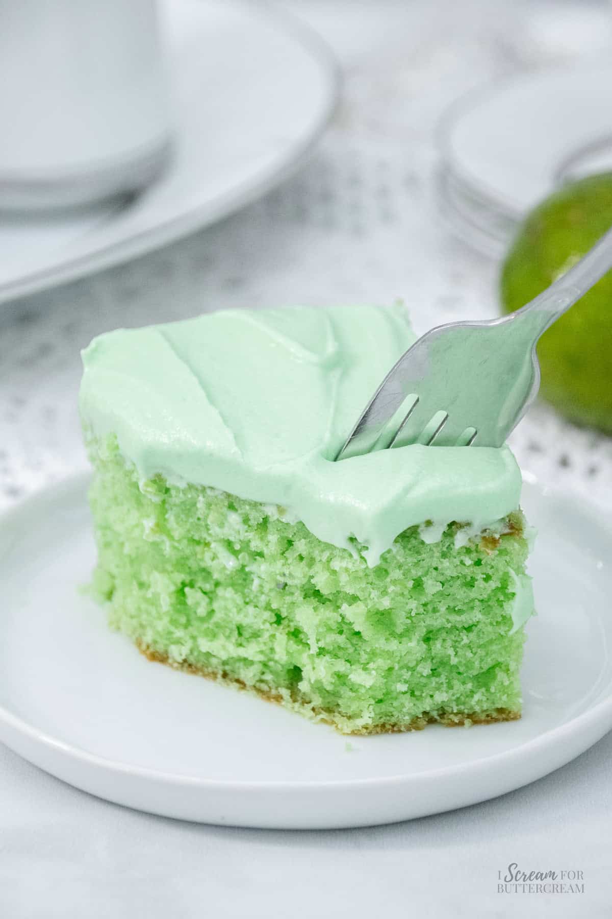 Fork cutting into a fluffy slice of key lime cake topped with creamy green frosting on a white plate.