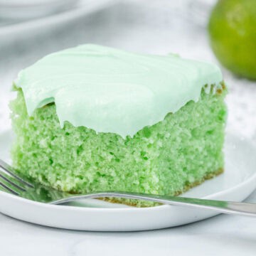 Slice of key lime cake with pale green frosting on a white plate, with a fresh lime in the background.