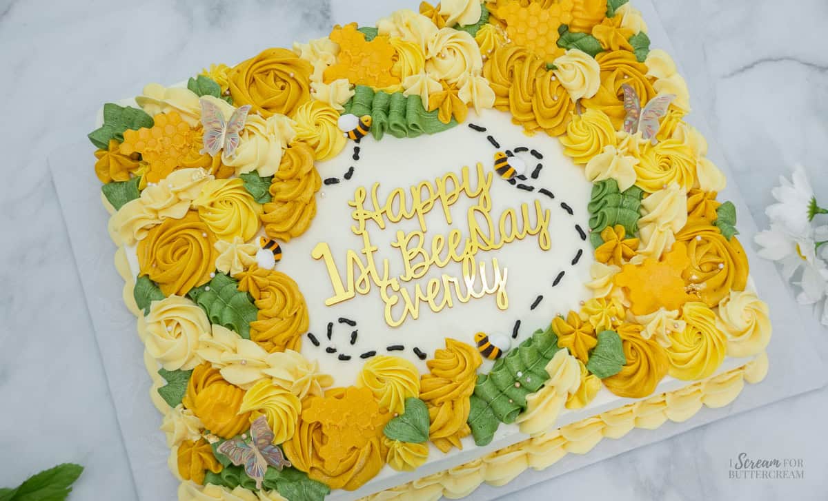 A rectangular sheet cake decorated with yellow, gold, and green buttercream flowers, fondant honeycombs, tiny fondant bees, and a gold