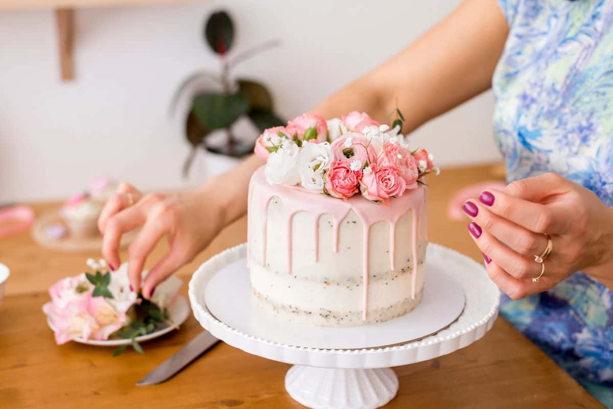 A person decorates a semi-naked cake with pink drip icing and fresh flowers, placing blooms on top of the cake while it sits on a white pedestal stand