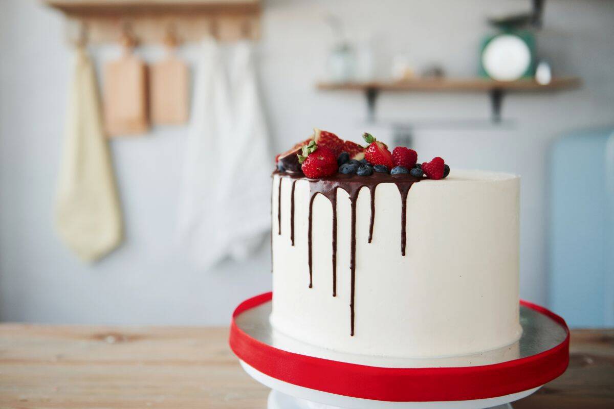 A white frosted cake with a glossy chocolate ganache drip, topped with fresh strawberries, blueberries, and raspberries, displayed on a cake stand with a red ribbon in a cozy kitchen setting.