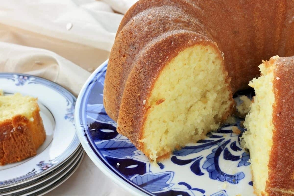 A golden, moist bundt cake with a slice cut out, served on a blue and white floral-patterned platter. A stack of matching plates with a slice of the cake sits nearby on a cream-colored fabric backdrop.