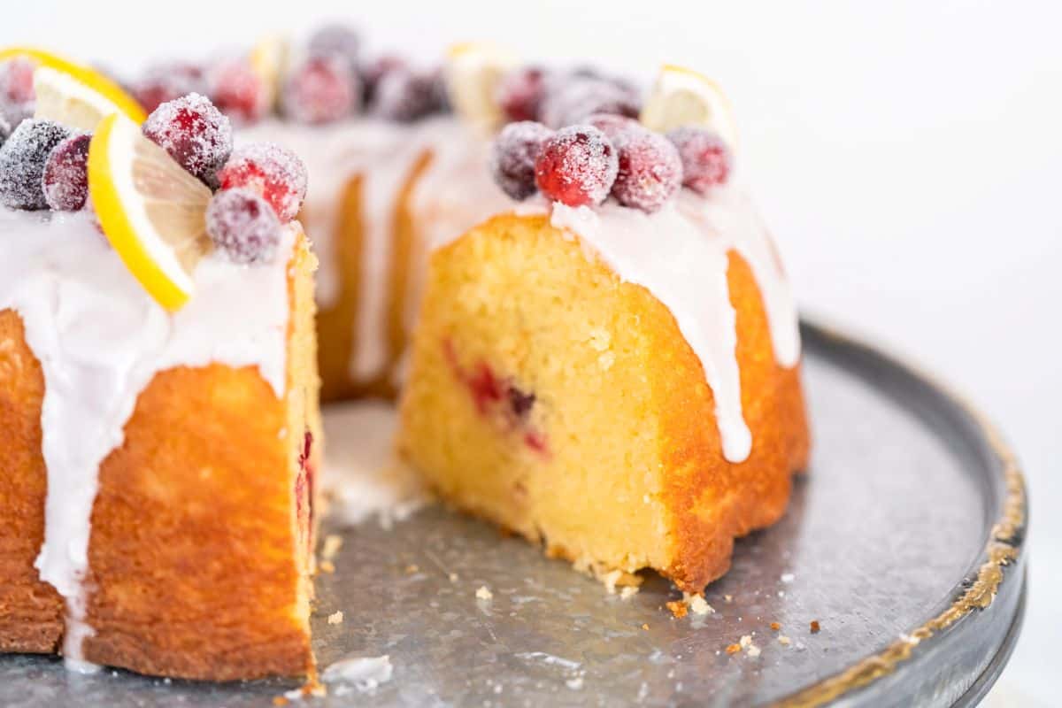 A sliced bundt cake topped with white icing, sugared cranberries, and lemon slices, displayed on a silver cake stand.