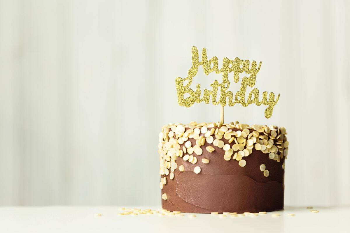 A chocolate frosted birthday cake decorated with gold confetti sprinkles and topped with a glittery gold "Happy Birthday" cake topper, set against a soft white background.