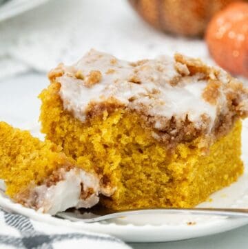 A thick slice of pumpkin coffee cake with a cinnamon streusel topping and vanilla glaze, served on a white plate with a fork and fall-themed decorations in the background.