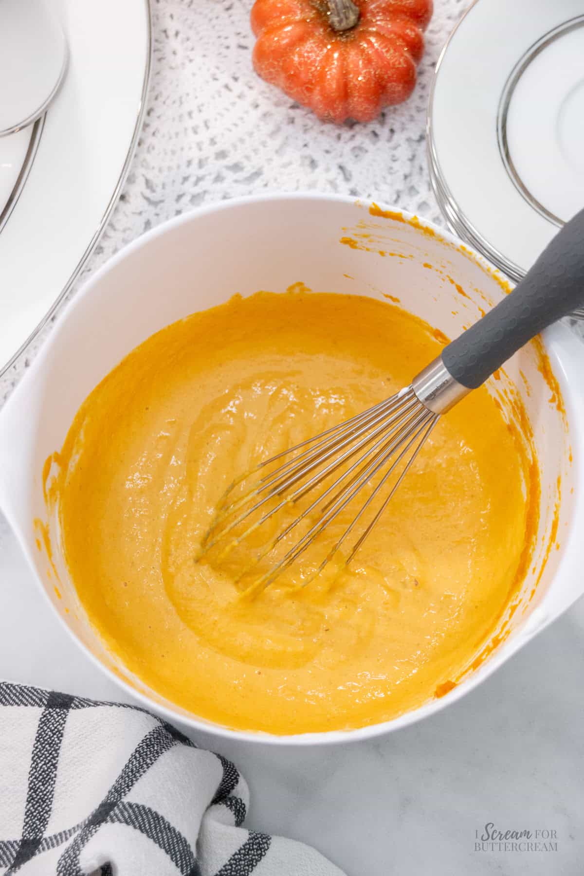 Pumpkin cake batter being mixed in a white bowl with a whisk, set on a light surface with fall decor and stacked plates in the background.