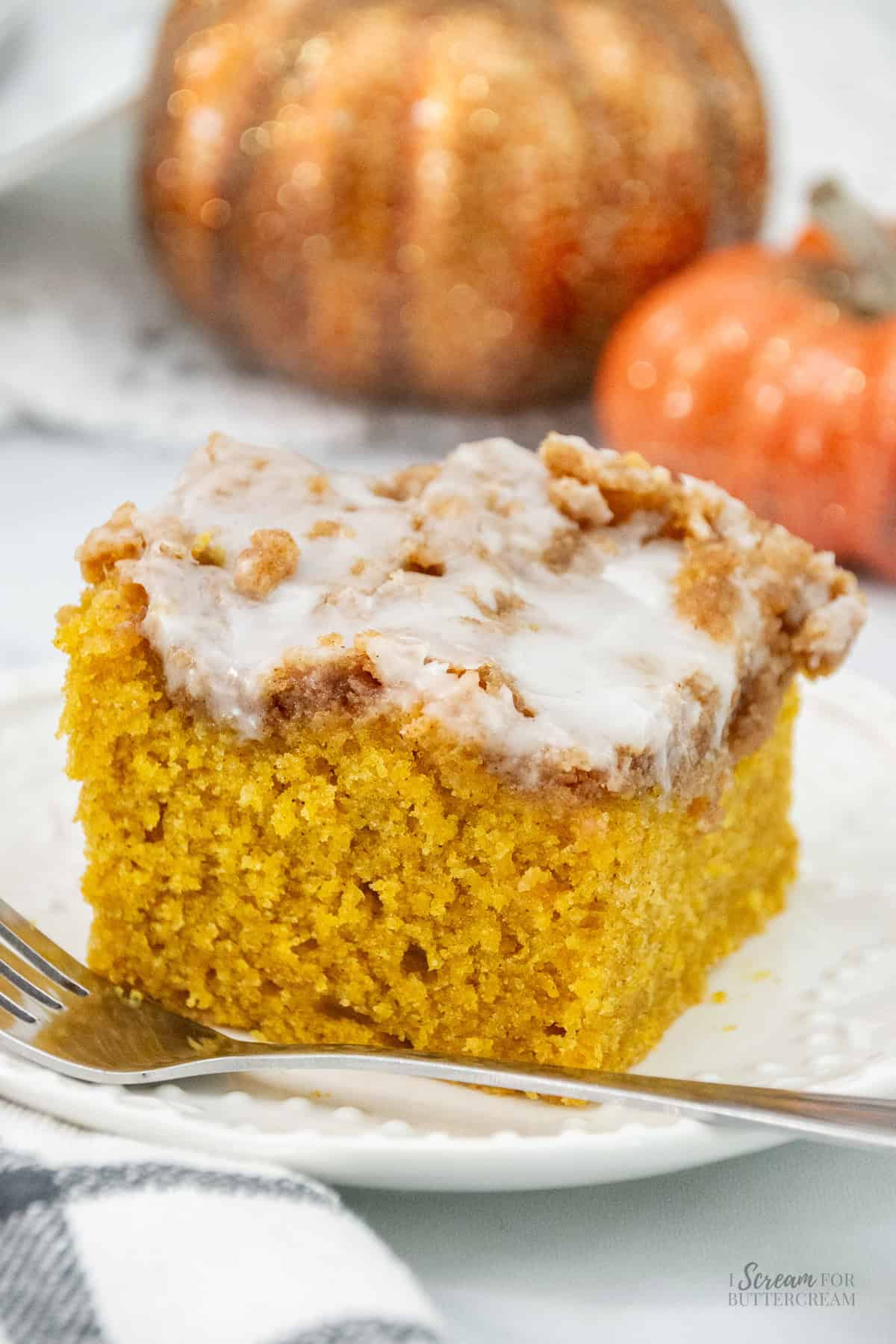 A single slice of moist pumpkin coffee cake topped with cinnamon streusel and icing, served on a white plate with a fork, with glittery decorative pumpkins in the soft-focus background.
