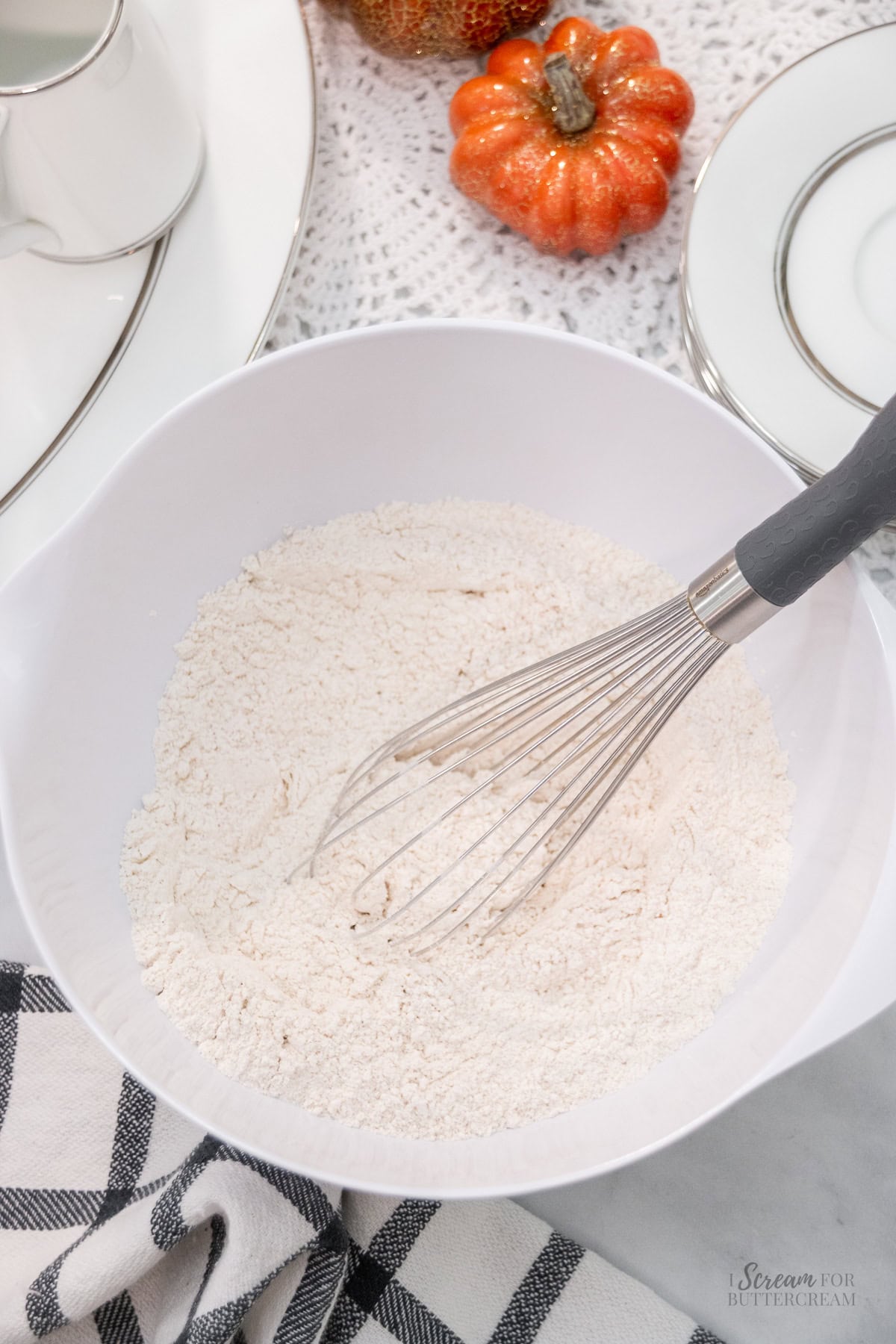 White mixing bowl filled with dry ingredients and a whisk resting inside, surrounded by fall-themed decor and stacked plates on a lace tablecloth.