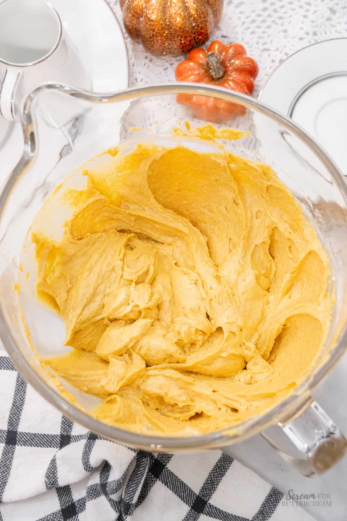 Thick pumpkin cake batter in a clear glass mixing bowl, resting on a white surface with a black checkered towel and fall-themed pumpkin decor in the background.