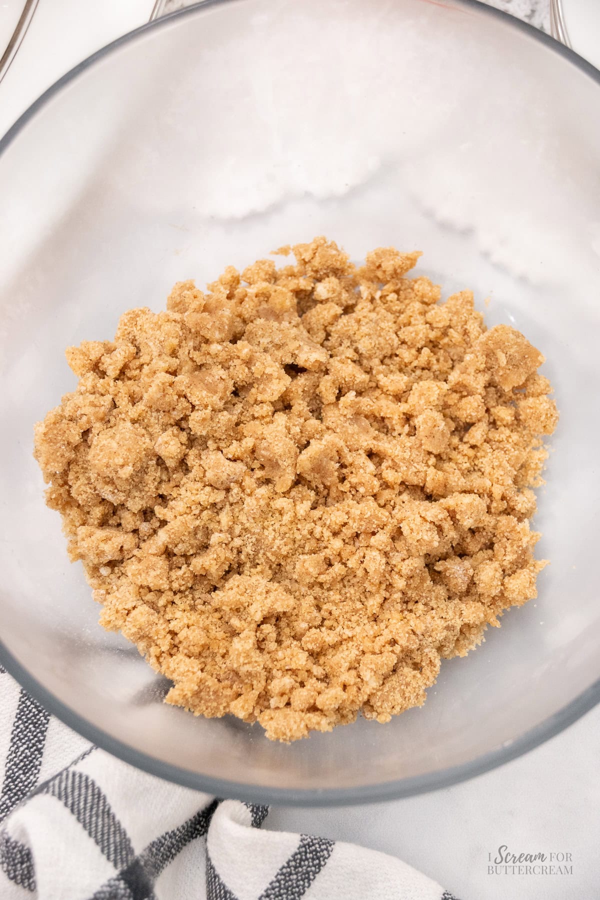 Bowl of crumbly cinnamon streusel topping in a clear glass bowl, resting on a white surface with a black-and-white checkered towel nearby.