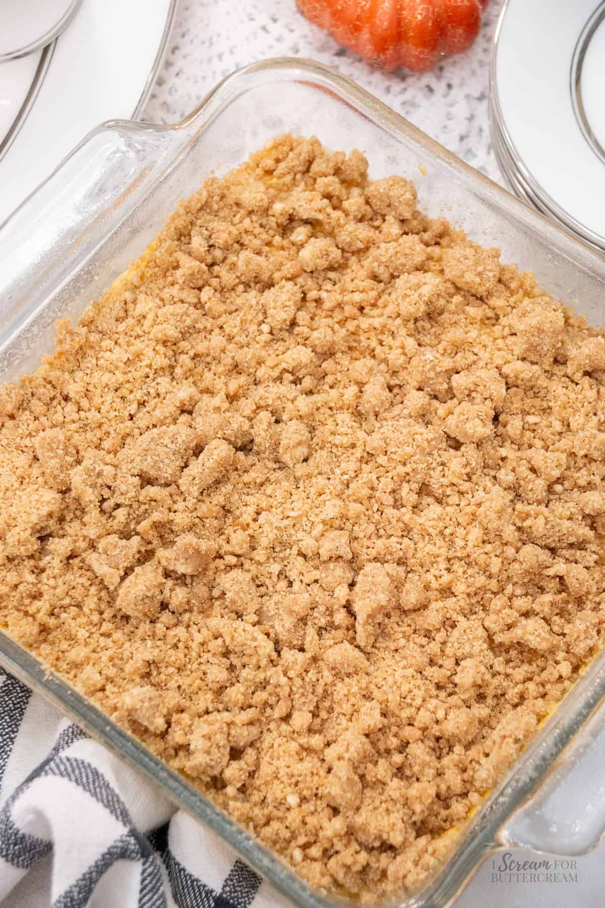 Unbaked pumpkin coffee cake in a glass baking dish topped with a thick layer of cinnamon streusel, ready to go in the oven with fall-themed decor in the background.