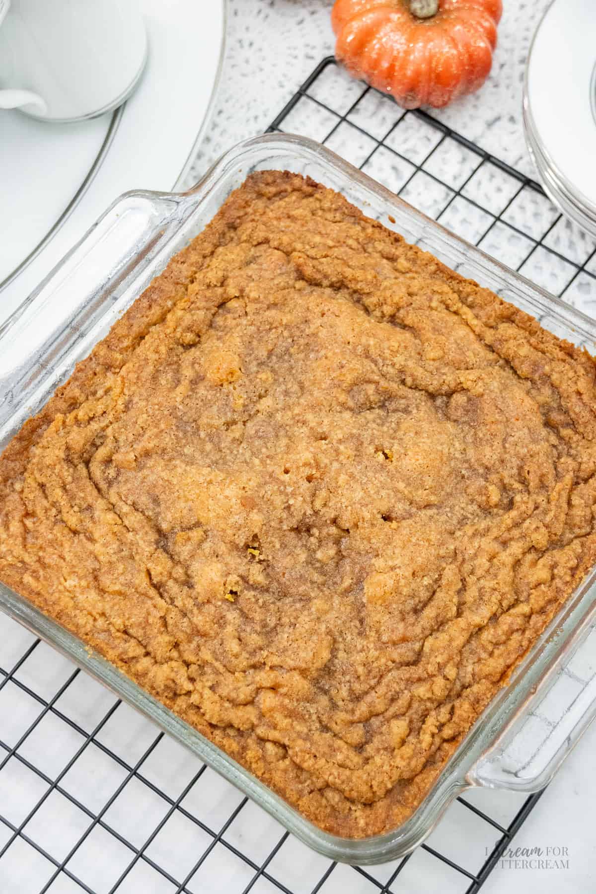 Freshly baked pumpkin coffee cake in a glass dish with a golden-brown cinnamon streusel top, cooling on a black wire rack with fall decor in the background.