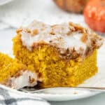 Close-up of a moist pumpkin coffee cake slice topped with cinnamon streusel and vanilla glaze, served on a white plate with a fork, with decorative pumpkins in the background.