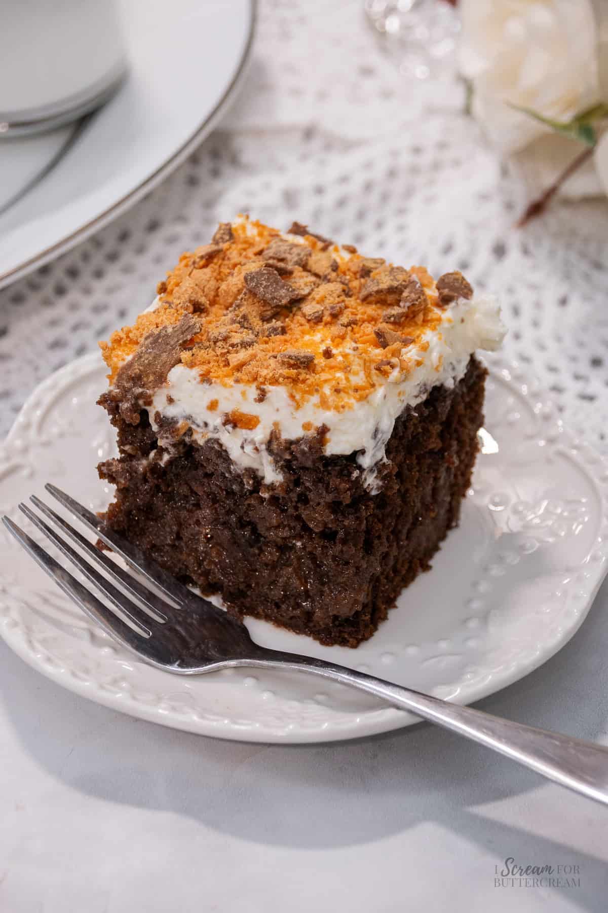 Slice of chocolate poke cake topped with whipped cream and crushed Butterfinger, served on a white plate with a fork.
