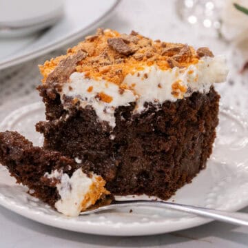 Close-up of a rich chocolate poke cake slice with whipped topping and crushed Butterfinger pieces, served on a white plate with a fork.