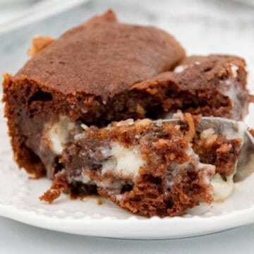 Close-up of a gooey chocolate earthquake cake slice with creamy filling on a white plate, with a spoonful taken out.