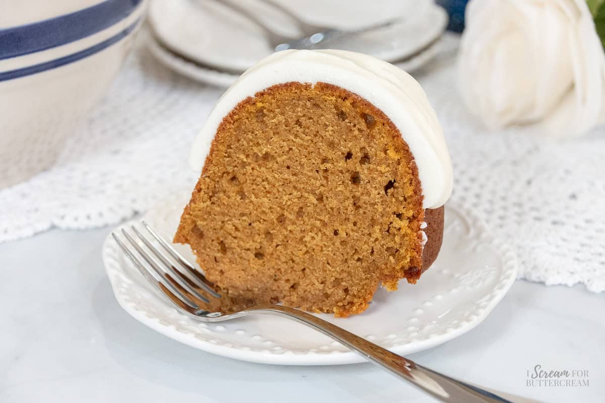 Slice of moist pumpkin spice Bundt cake topped with thick cream cheese glaze on a white dessert plate with a fork.